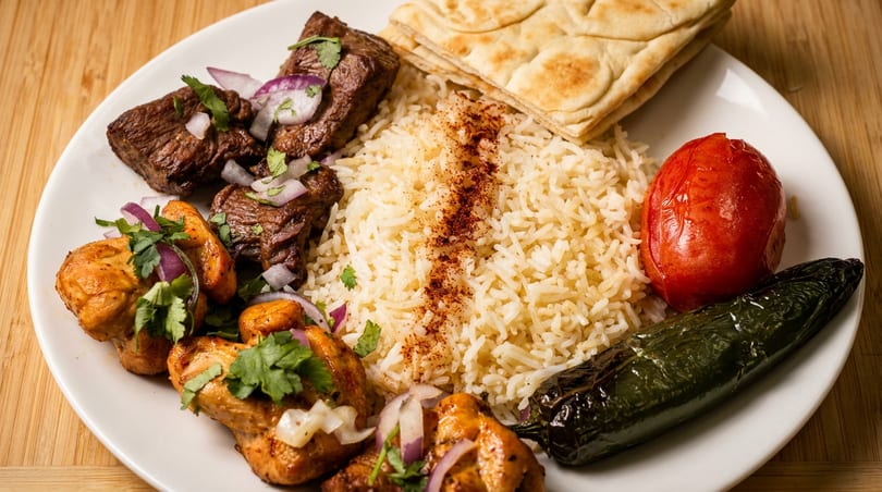 A plate of chicken and beef kebabs with rice, flatbread, grilled tomato, and jalapeño.