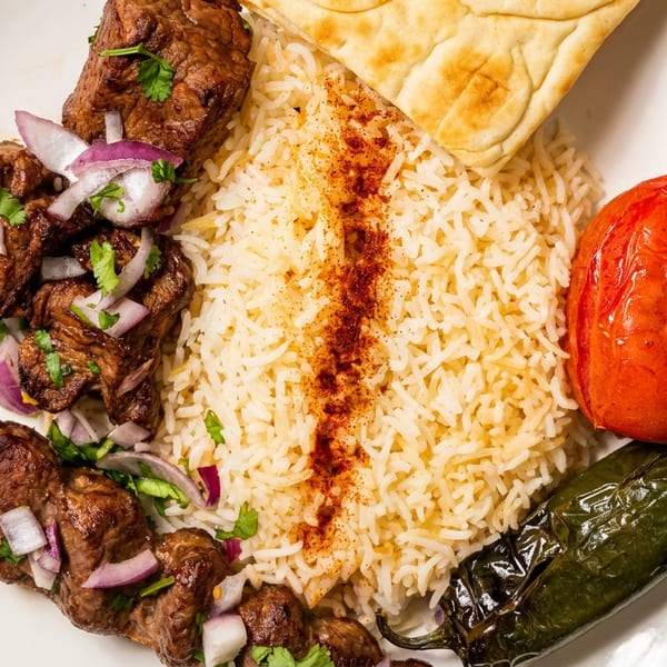 Grilled meat skewers with rice, roasted tomato, green pepper, and flatbread on a white plate.