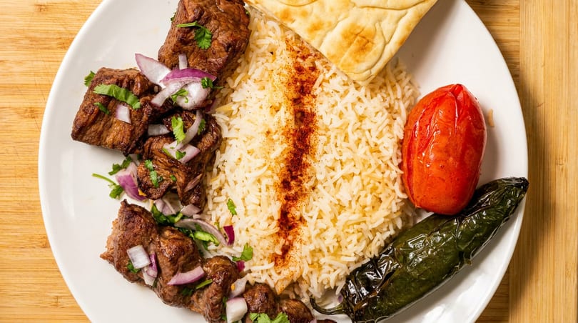 Grilled meat skewers with rice, roasted tomato, green pepper, and flatbread on a white plate.