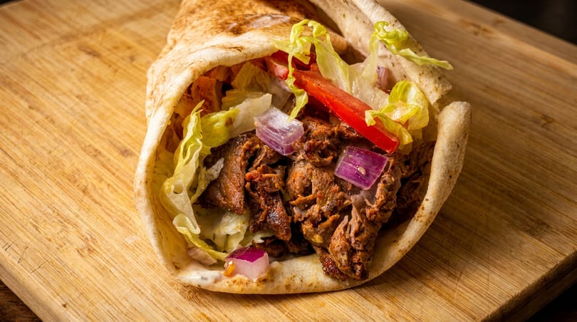 A close-up of a shawarma wrap filled with meat, lettuce, tomatoes, and red onions on a wooden board.