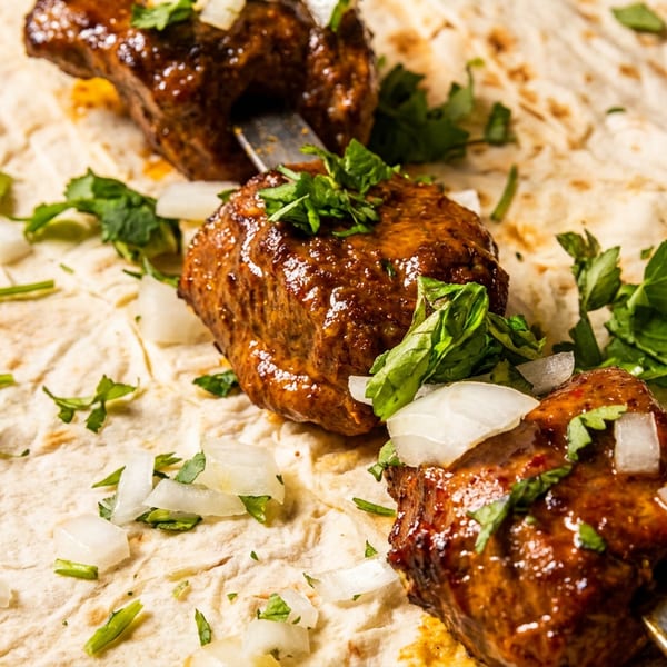 Three skewers of glistening meat kebabs, garnished with fresh herbs and diced onions, resting on flatbread.