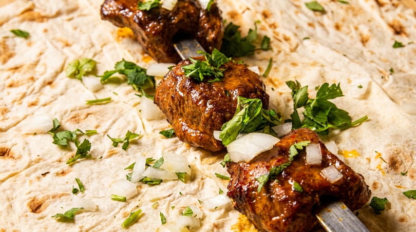 Three skewers of glistening meat kebabs, garnished with fresh herbs and diced onions, resting on flatbread.